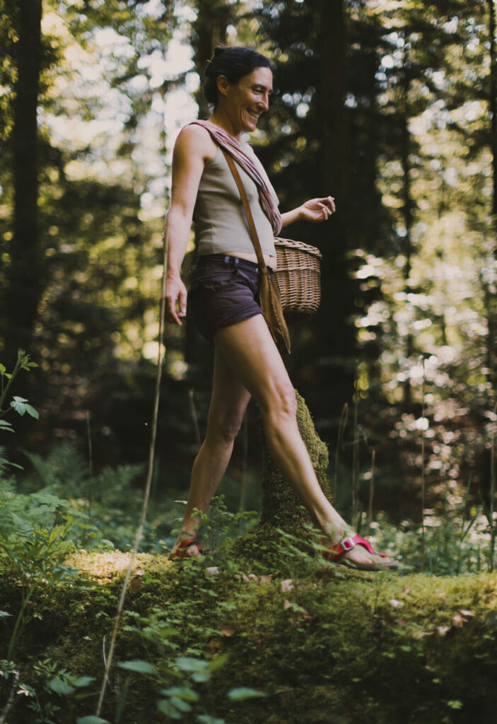 Sarah se balade dans une forêt luxuriante avec un grand sourire et tient son panier de cueillette à l'aide de son bras.