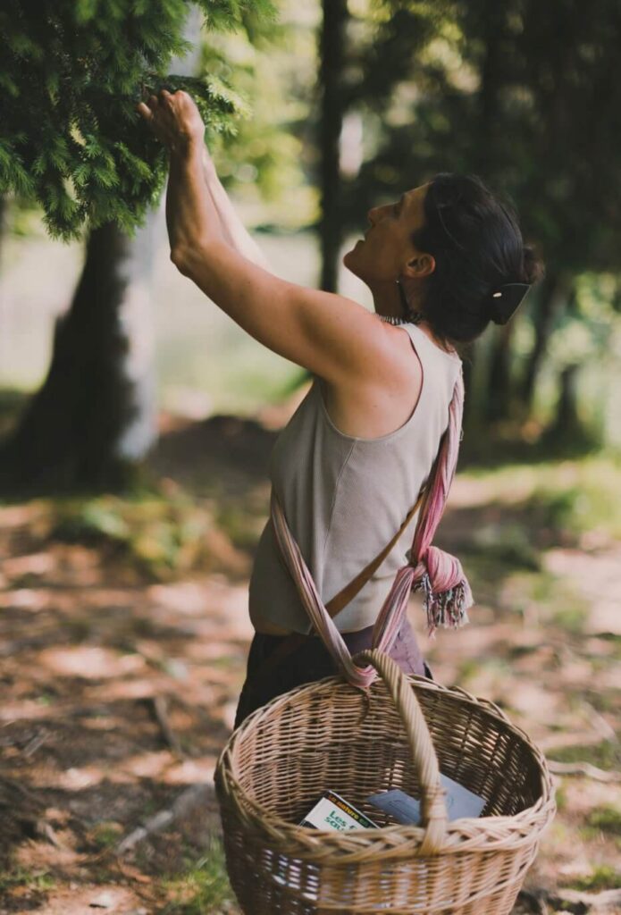 Sarah a ses bras en hauteur pour cueillir une plante et elle est accompagnée de son panier tenu en bandoulière.