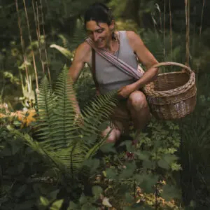 Souriante, Sarah est accroupie sur le sol, près d'une fougère et entourée d'herbes hautes.