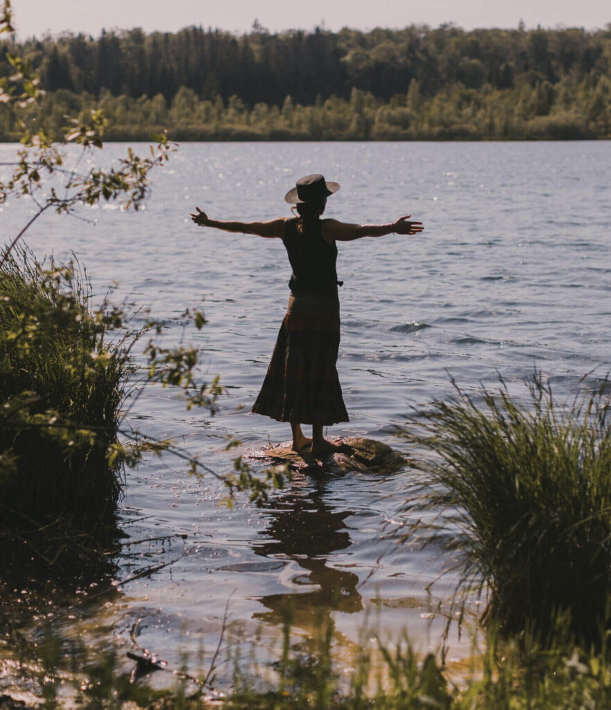 Au bord d'un lac du Jura et sous un ciel ensoleillé, Sarah se tient debout sur un rocher avec les bras grands ouverts.
